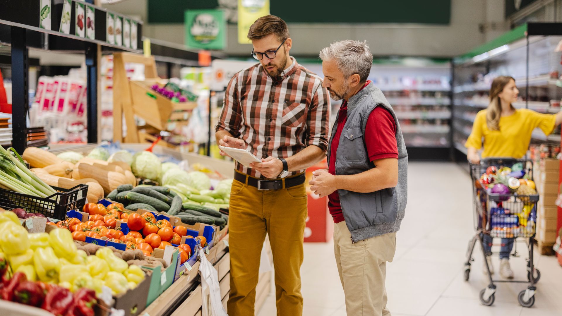 Two grocery store employees discussing inventory using a tablet in the produce section, surrounded by fresh vegetables and shoppers.