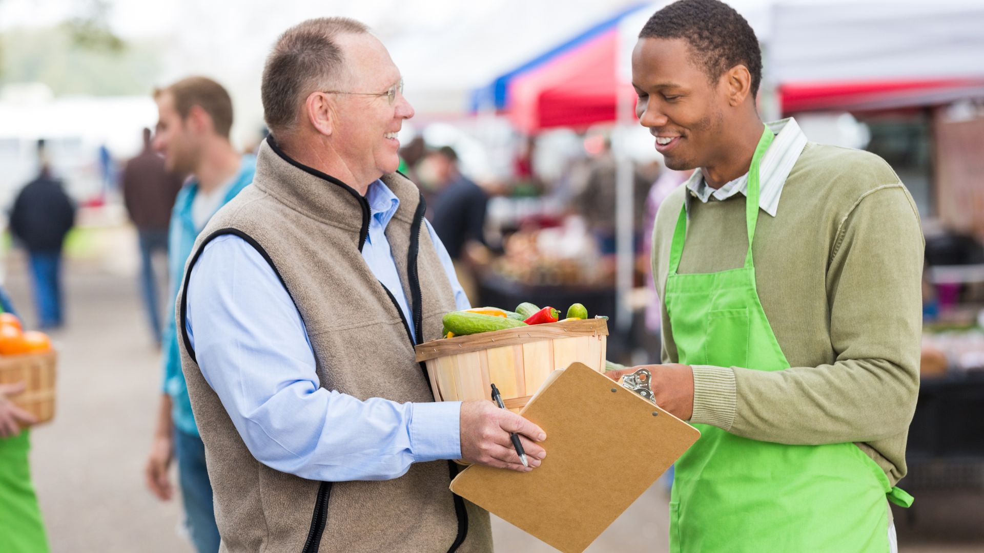 A farmers’ market manager in a green apron speaks with a smiling vendor holding a basket of fresh vegetables and a clipboard.