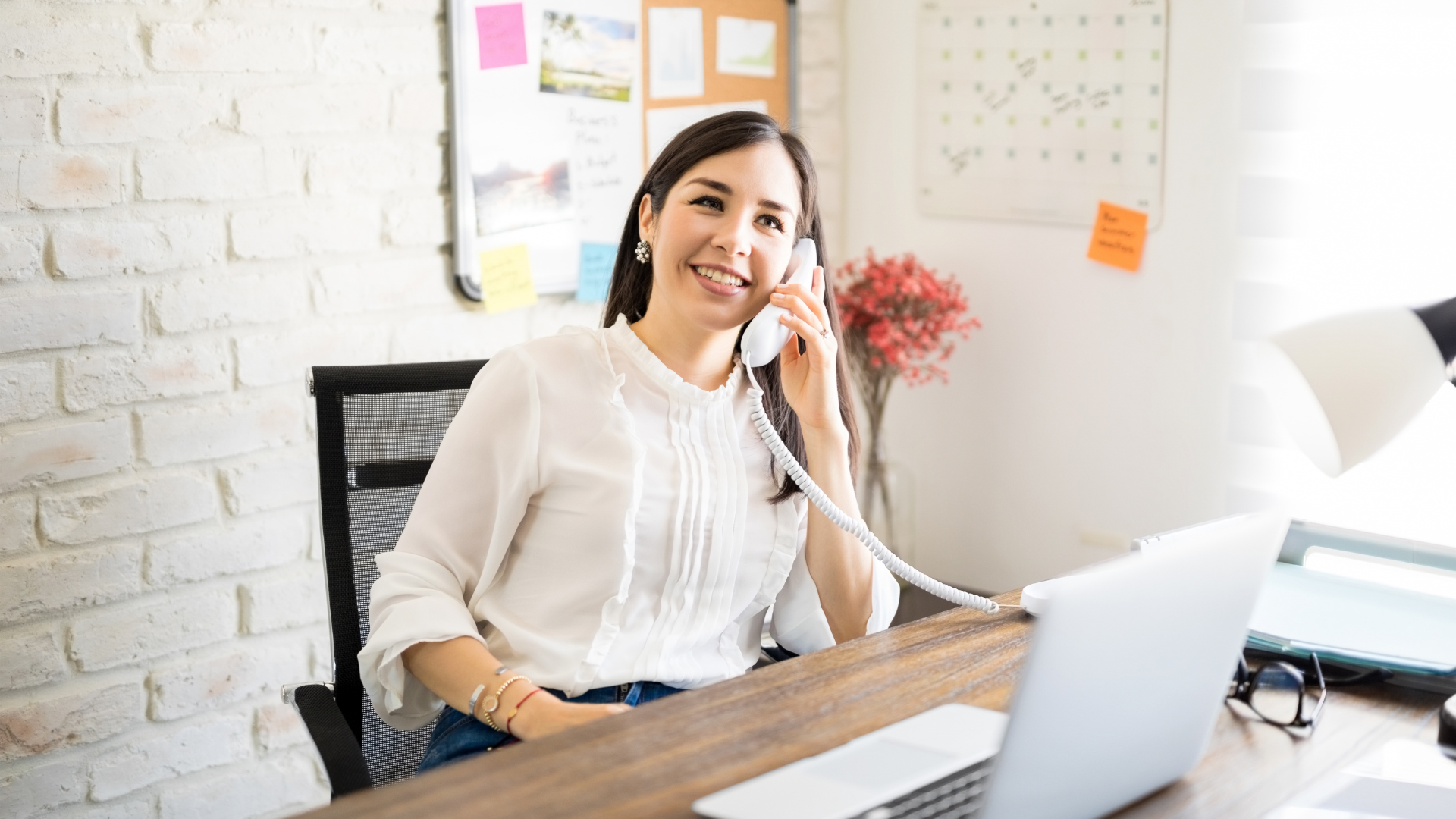 Smiling woman talking on a landline phone at her desk in a bright office, with a laptop, calendar, and bulletin board in the background.