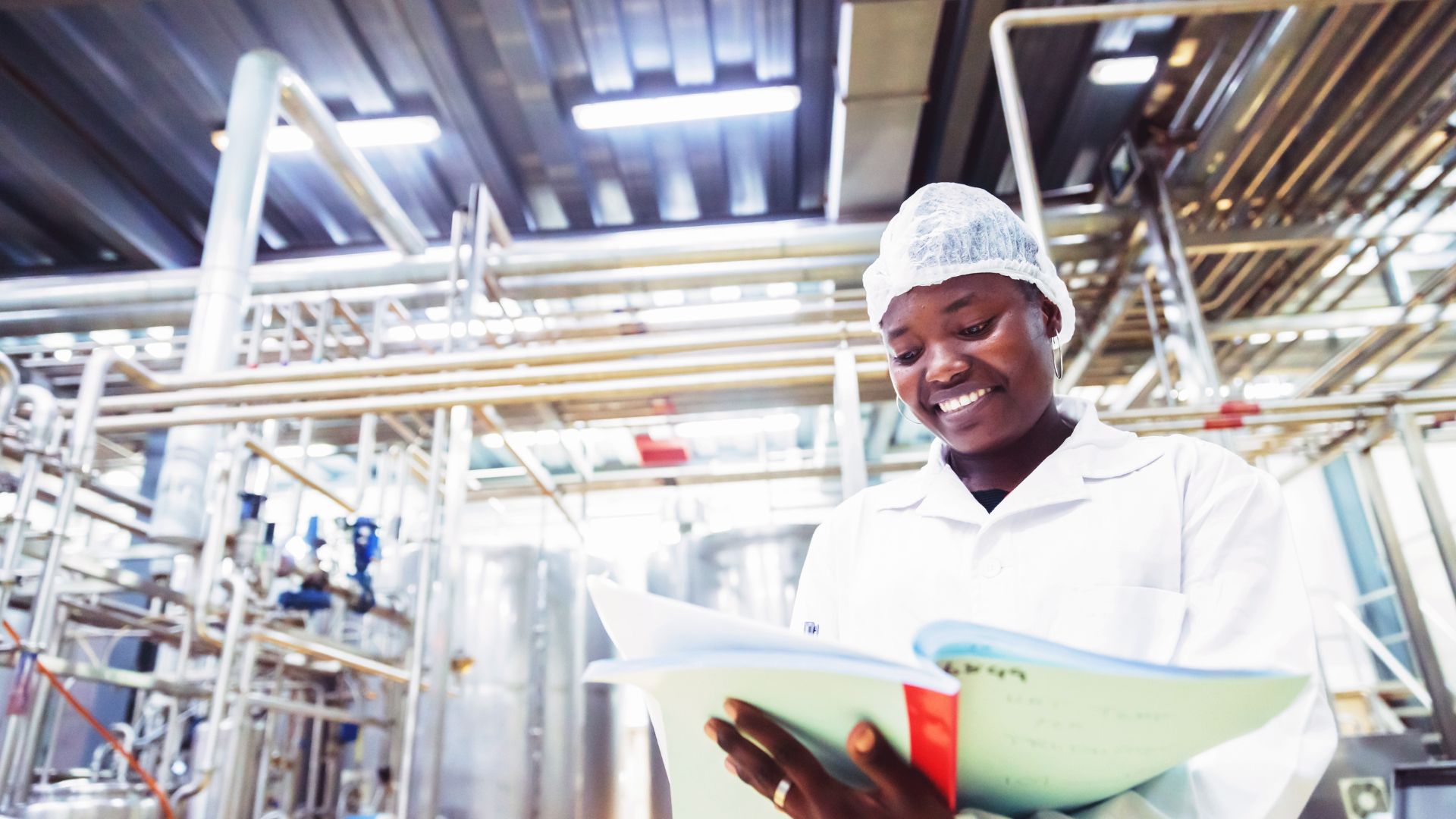 Food auditor in a lab coat and hairnet reviewing inspection notes inside a food processing facility, surrounded by stainless steel equipment and piping systems.