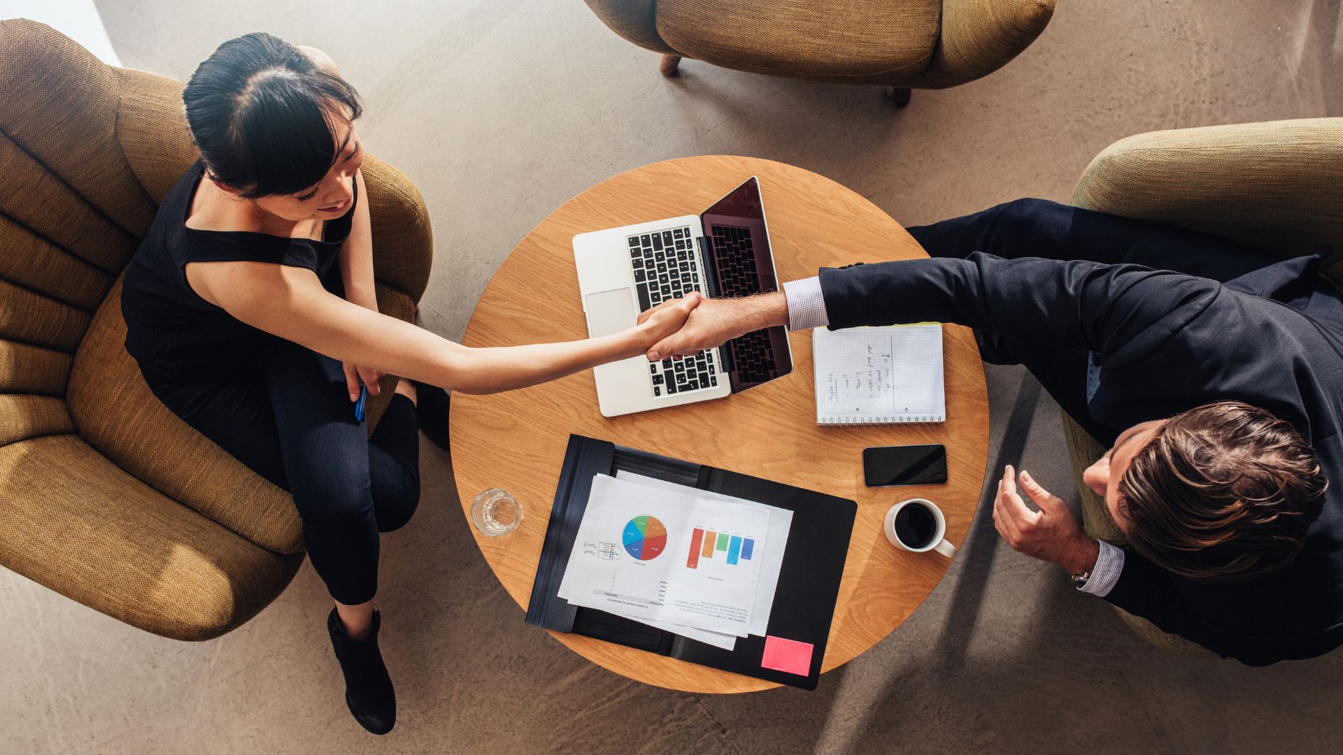 Food broker and client shaking hands at a meeting table with sales charts, a laptop, and product notes—finalizing a food distribution or retail deal.