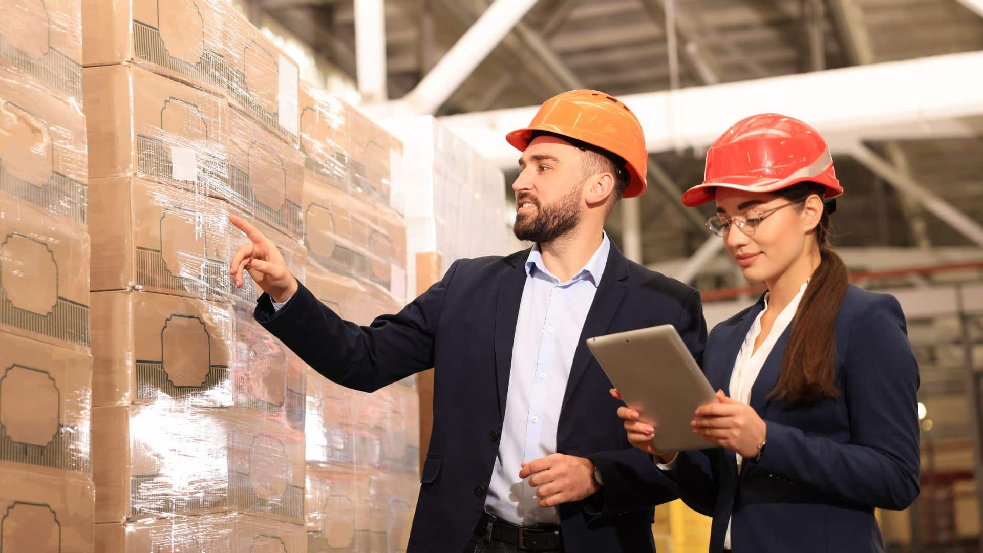 Two food production managers wearing safety helmets and business attire inspect stacked pallets in a warehouse, using a tablet and pointing at inventory.