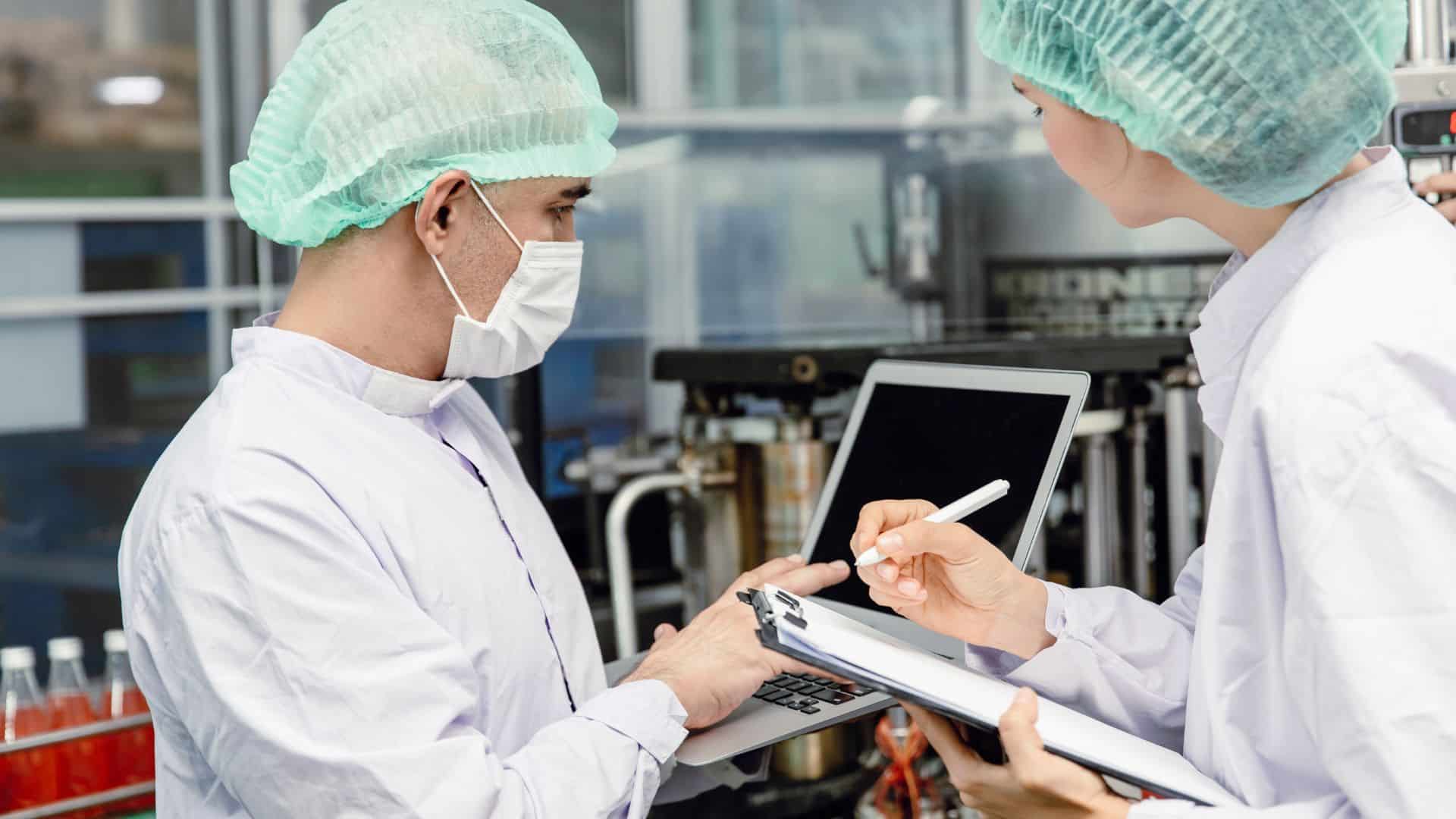 Two quality assurance technicians in protective gear inspecting food production data on a laptop and clipboard inside a manufacturing facility.