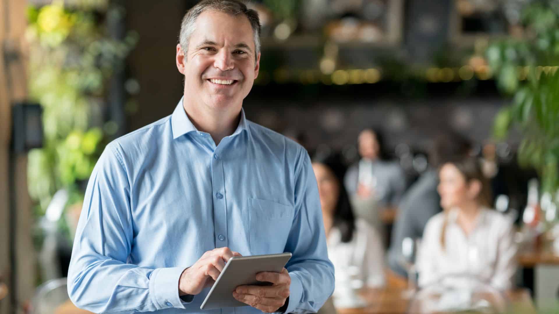Smiling restaurant manager holding a tablet while standing in a busy dining area with customers in the background.