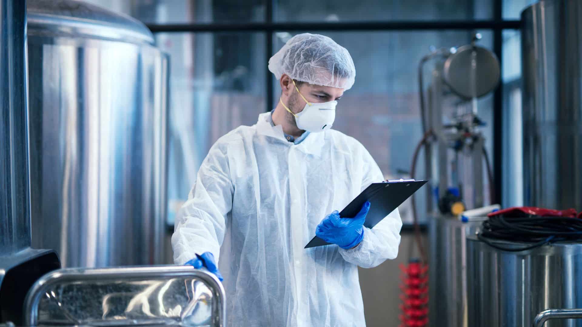 Sanitation manager wearing protective clothing and mask while inspecting industrial food processing equipment with a clipboard in a clean manufacturing facility.
