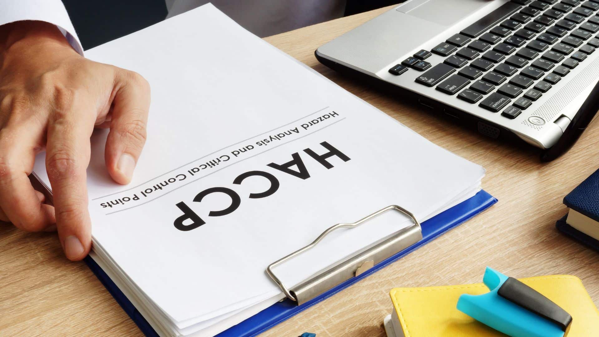 Close-up of a person reviewing a HACCP (Hazard Analysis and Critical Control Points) document on a clipboard next to a laptop, representing food safety compliance work.