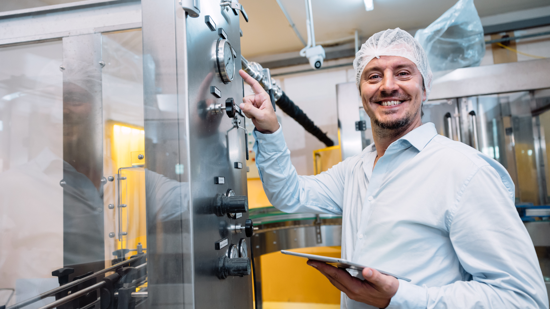 Machine operator in a food manufacturing facility adjusting equipment settings while holding a digital tablet, wearing a hairnet and smiling confidently.