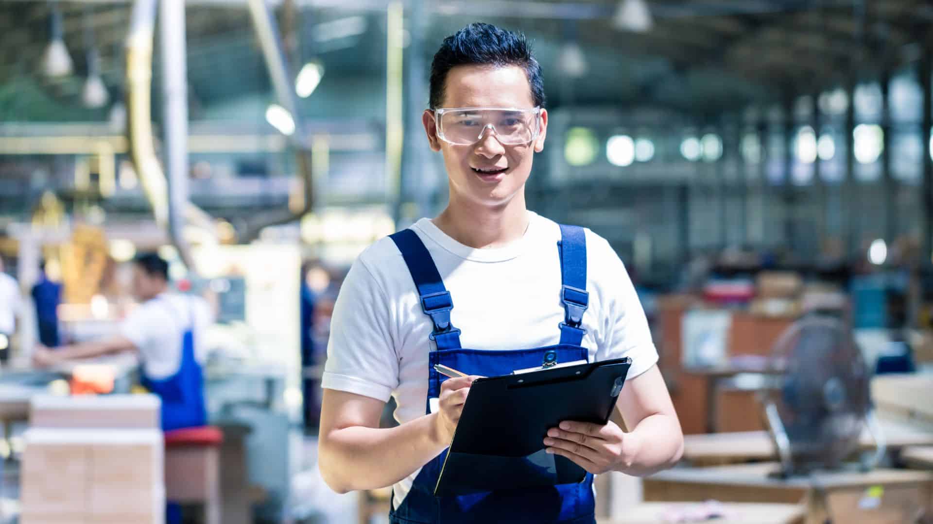 Production lead wearing safety goggles and overalls holds a clipboard while supervising operations in a food manufacturing facility.
