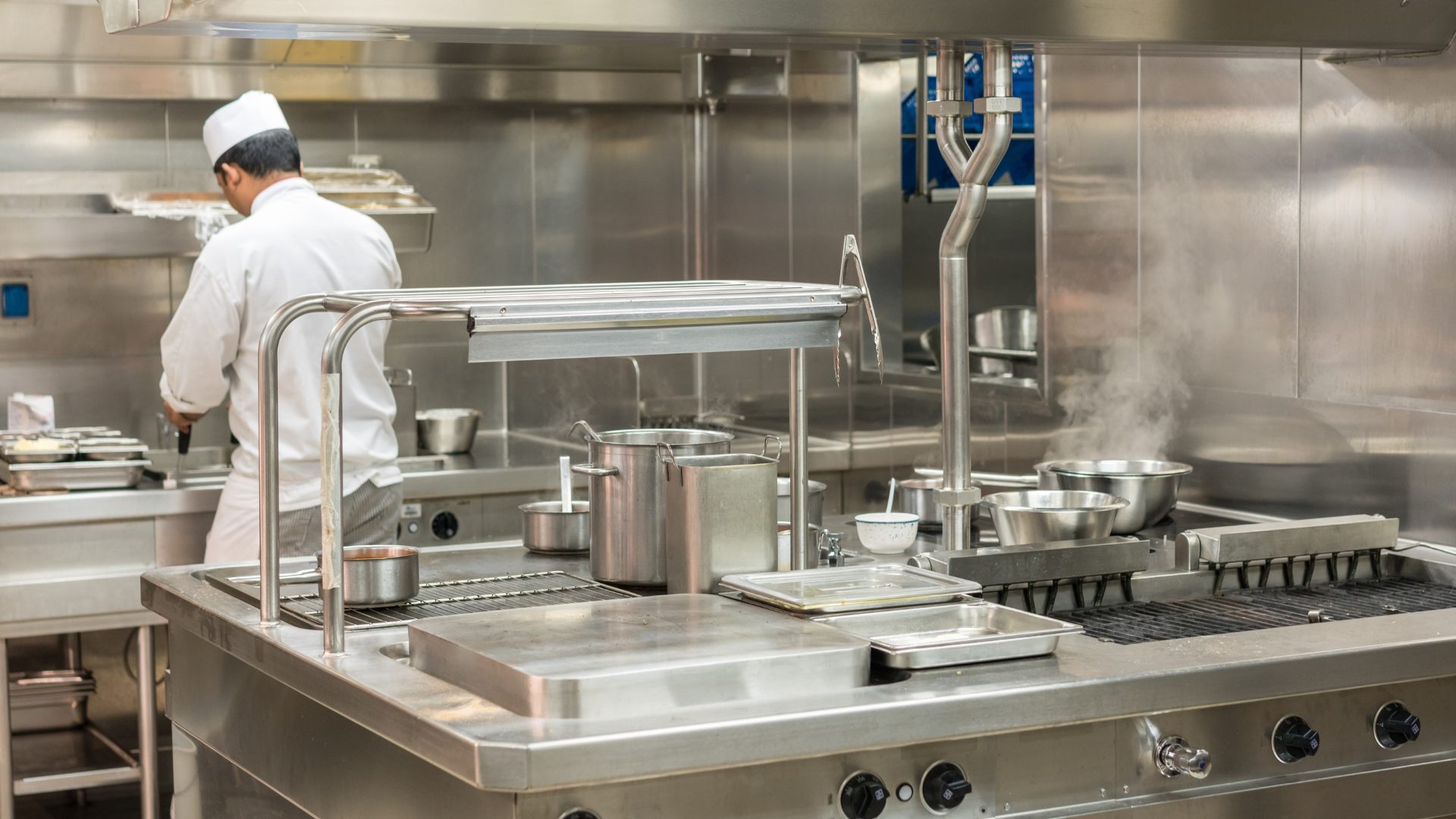 Research chef working in a commercial kitchen surrounded by stainless steel equipment and cooking pots, preparing ingredients at a stovetop.