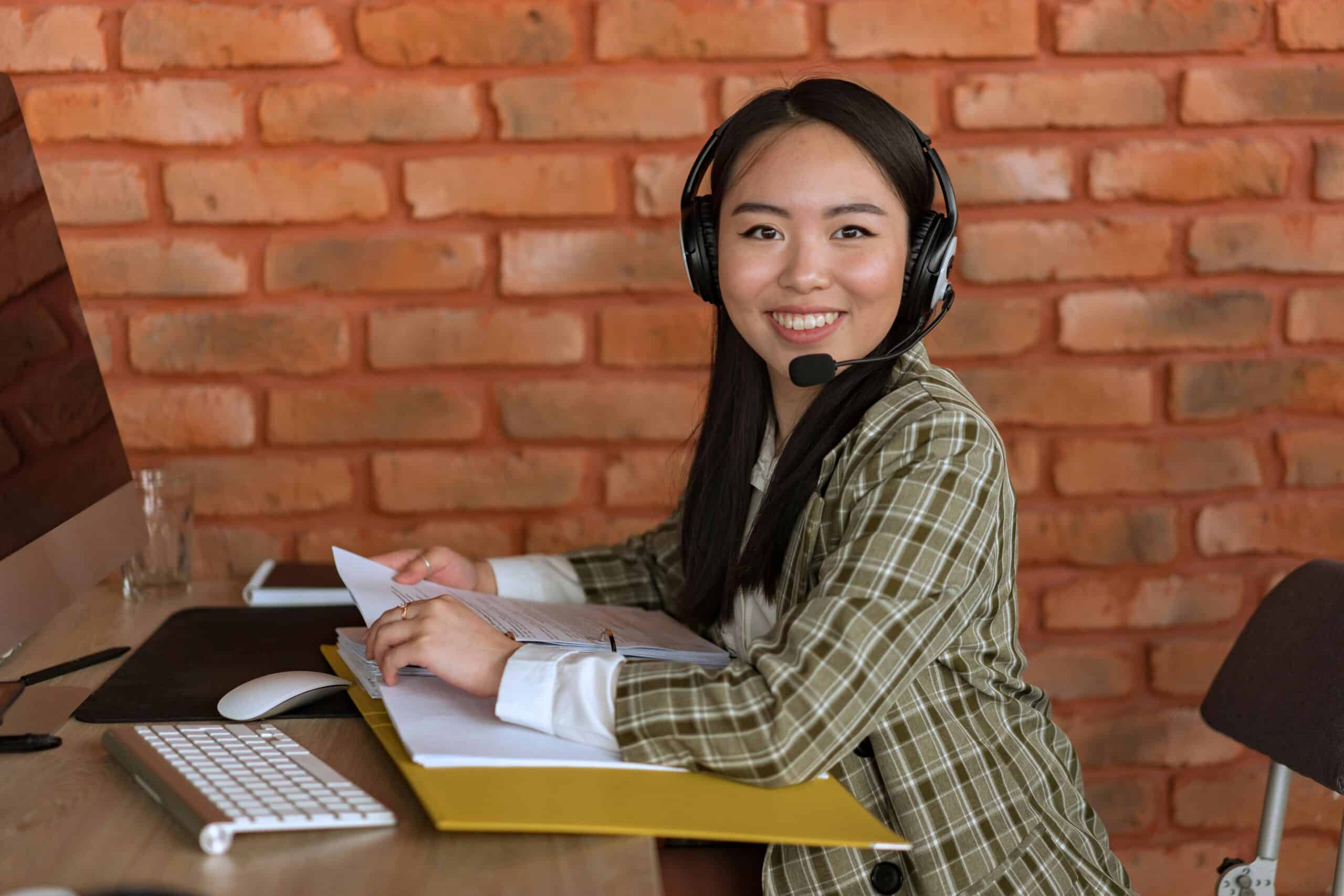 Smiling technical sales representative wearing a headset, working at a desk with documents and a computer in front of a brick wall background.