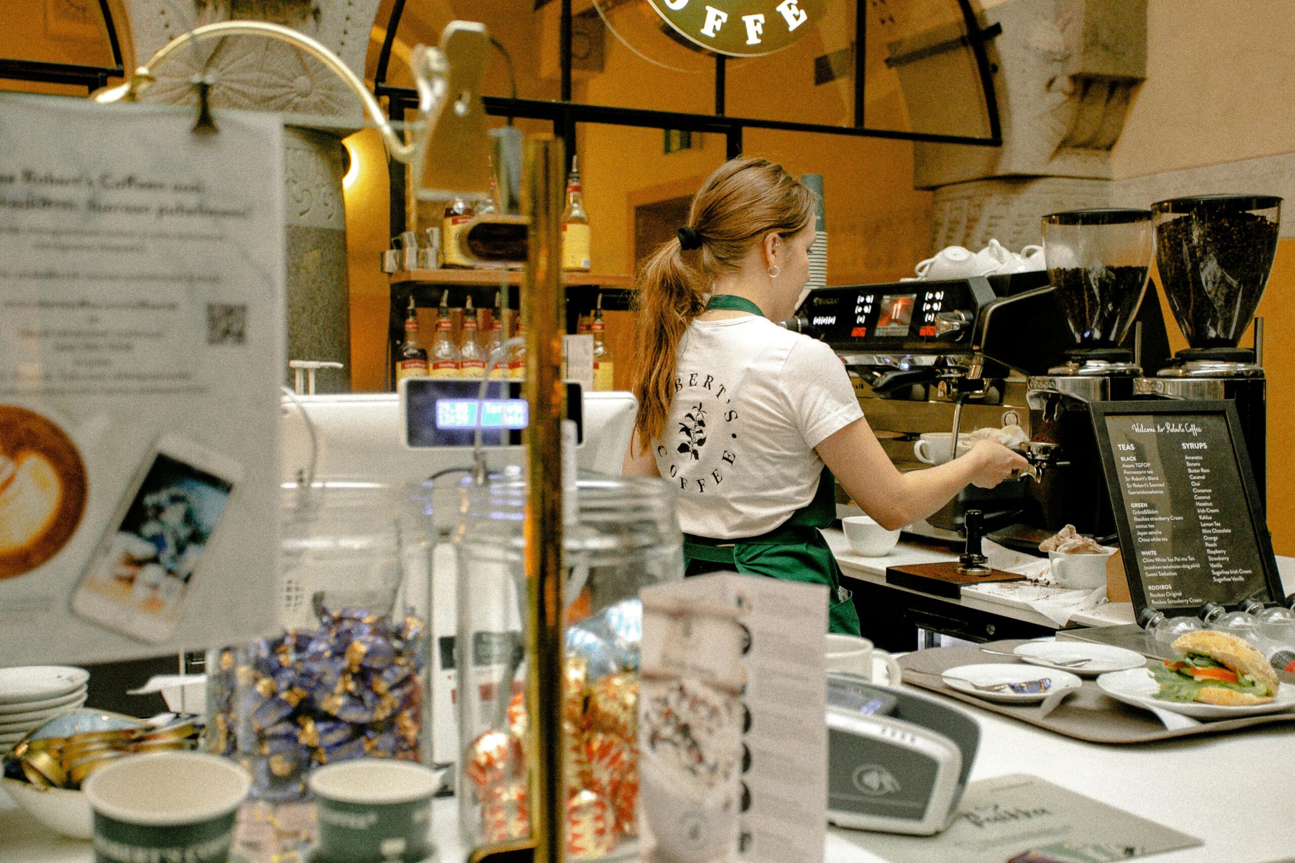 Barista preparing coffee behind a café counter with an espresso machine, menu board, and various syrups and snacks on display.