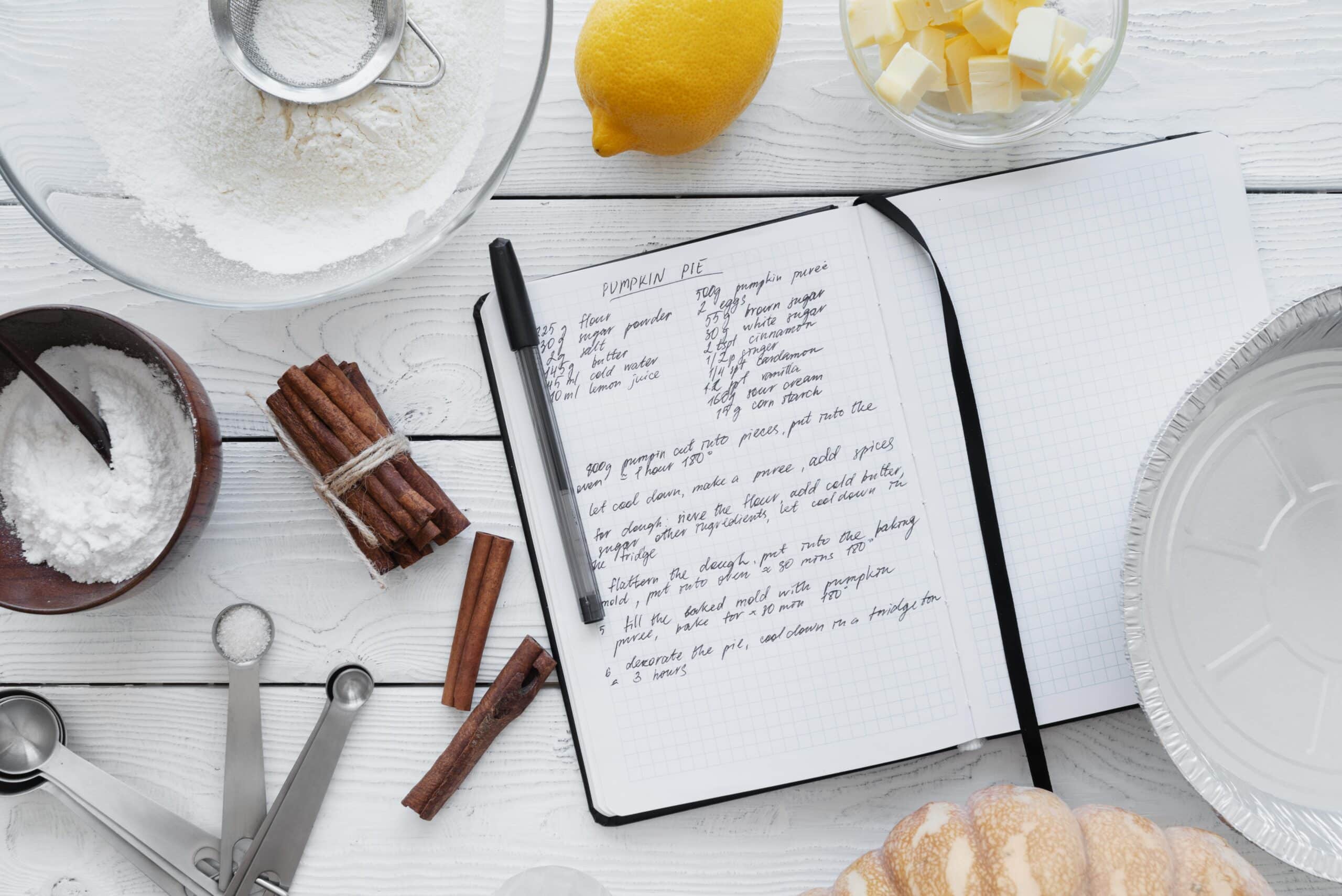 Top view of an open recipe book with a pumpkin pie recipe on a white wooden table, surrounded by ingredients and tools. A food science-focused recipe with precise measurements and ingredient interactions to enhance texture and flavor